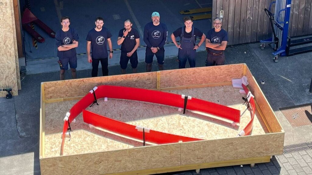 Eine Gruppe von sechs Personen in Arbeitskleidung steht vor einem großen Holzrahmen mit einer roten Konstruktion.
