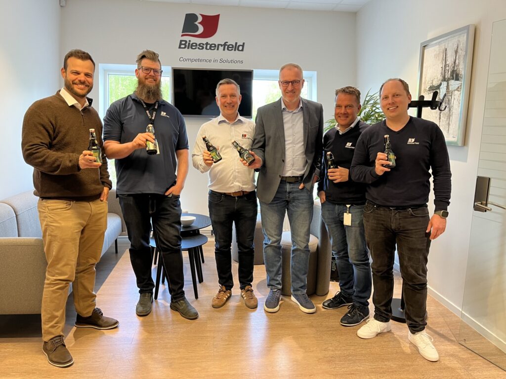 Six men are holding beer bottles in a modern office setting.